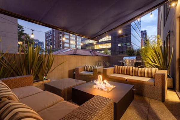 An outdoor patio seating area with sofas arranged around a fire pit under a covered terrace at dusk.