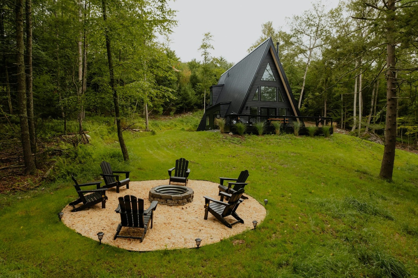 An A-frame sauna in a forest clearing with a circular fire pit and chairs in the foreground.