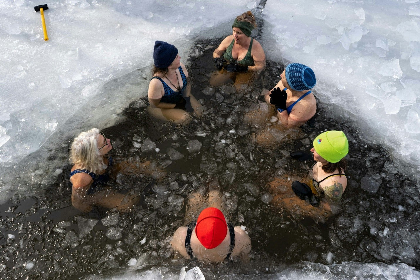 A group of people sitting in an icy water hole surrounded by snow and ice.